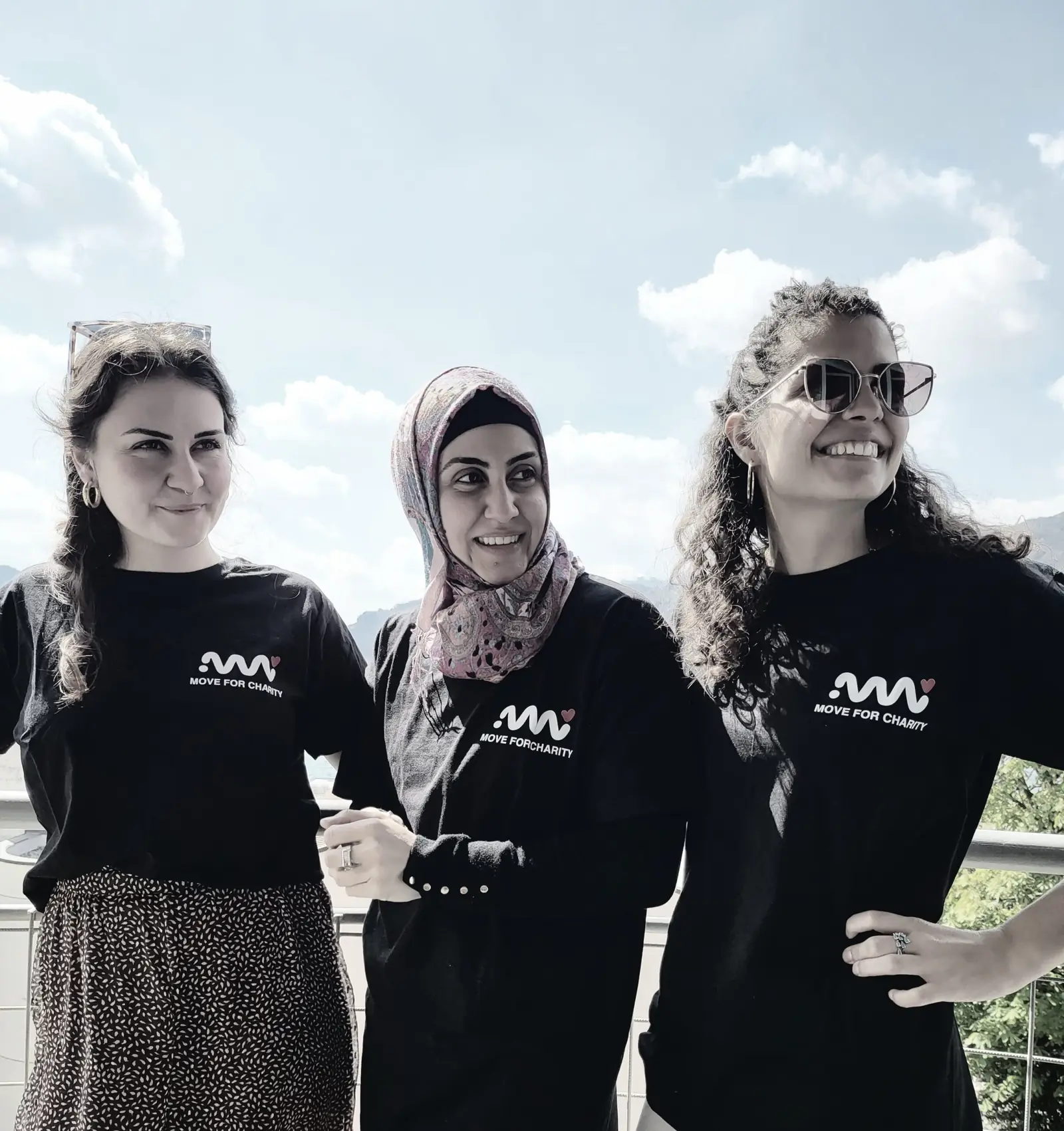 Tre volontarie con la maglietta Move for Charity posano su una terrazza con le montagne alle spalle, foto in bianco e nero