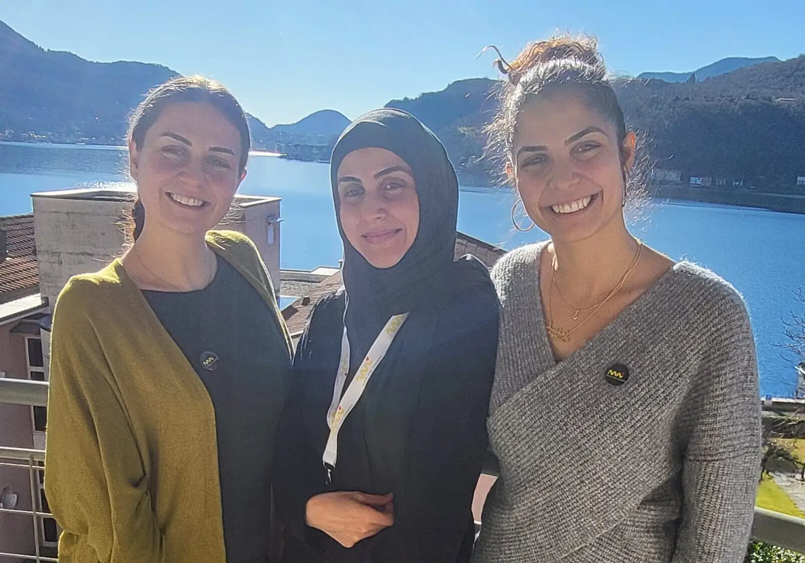 Tre donne sorridenti con la spilla di Move for Charity posano su una terrazza affacciata sul lago e sulle montagne