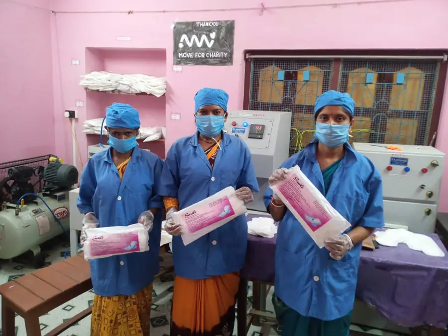 Tre operatrici in camice azzurro e mascherina mostrano le confezioni di assorbenti prodotte nel laboratorio, sotto lo striscione di ringraziamento a Move for Charity